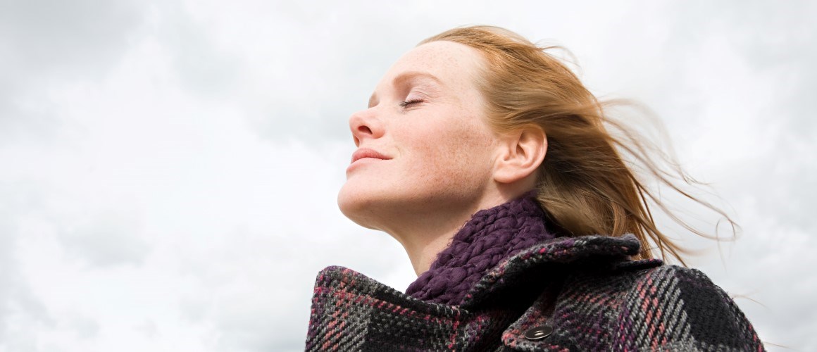 Woman with eyes closed, standing outside with a coat on, with wind blowing in her hair.