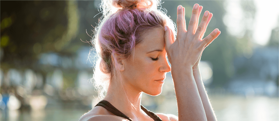 A woman in the park with her thumbs touching her forehead.