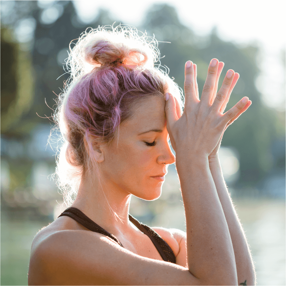 A woman in the park with her thumbs touching her forehead.