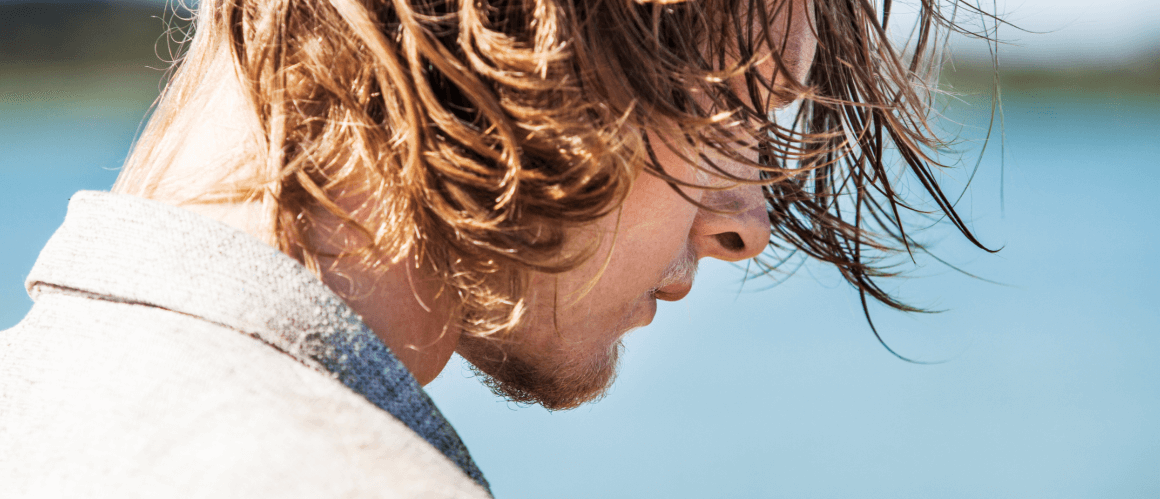 Man with long hair, standing in front of a body of water, with wind blowing in his hair.