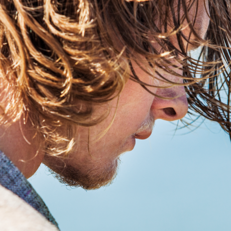 Man with long hair, standing in front of a body of water, with wind blowing in his hair.