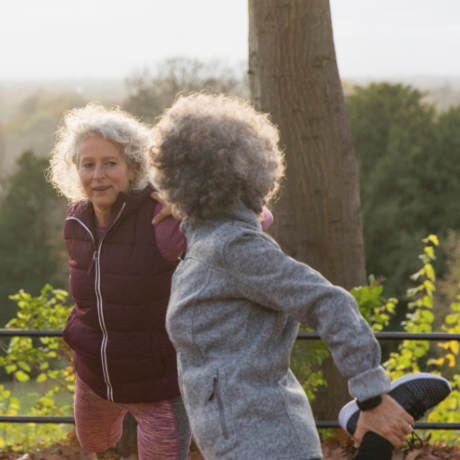 Two women wearing exercise attire, stretching and talking outdoors in a park.