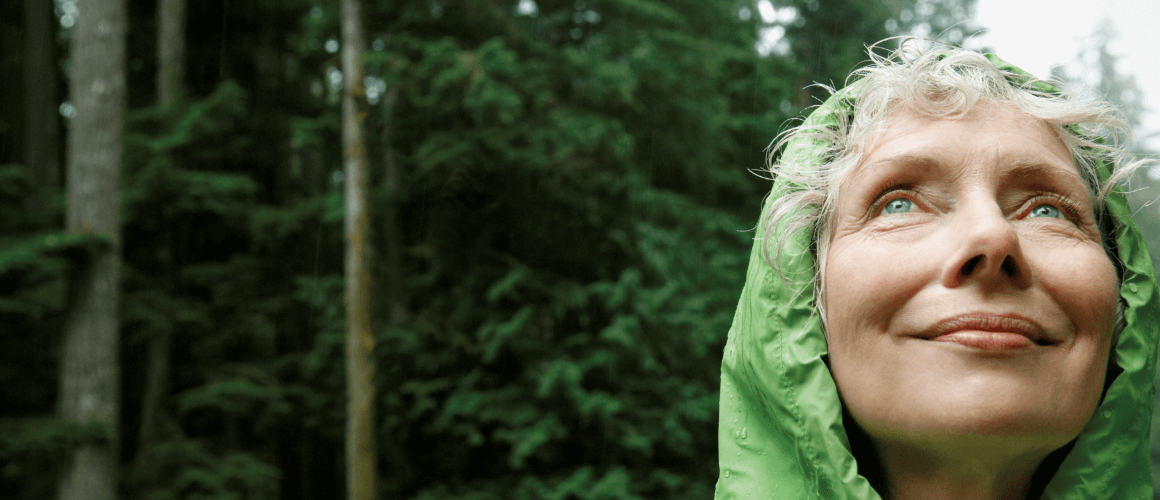 Woman wearing a green rain jacket, standing in a forest and looking up at the sky.