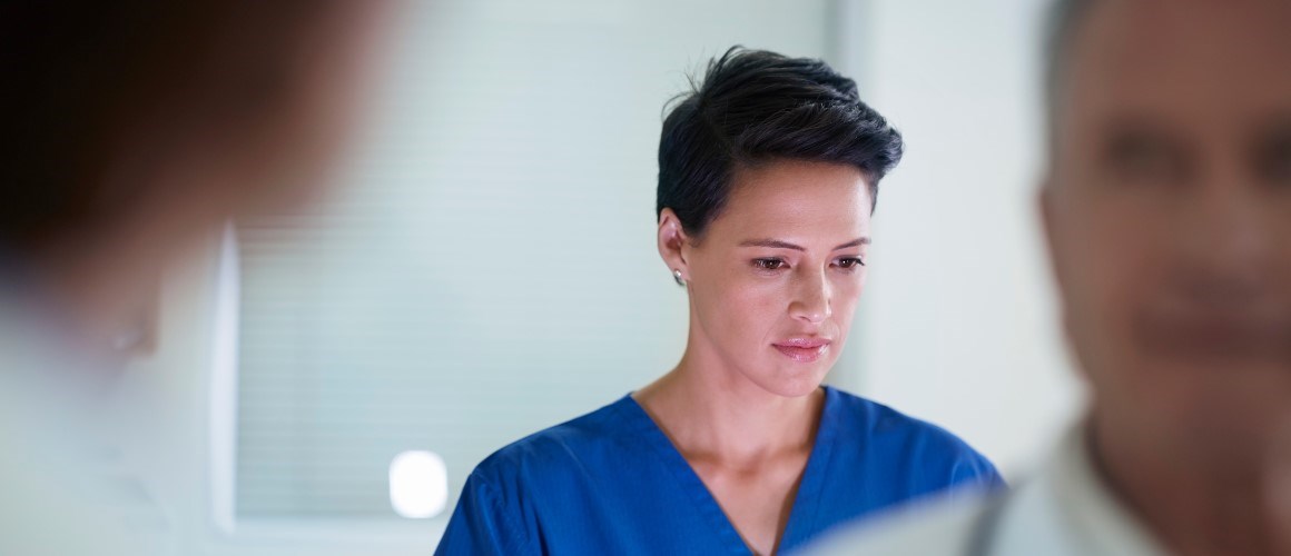 Female doctor wearing blue scrubs looking at computer with other doctors in the room. 