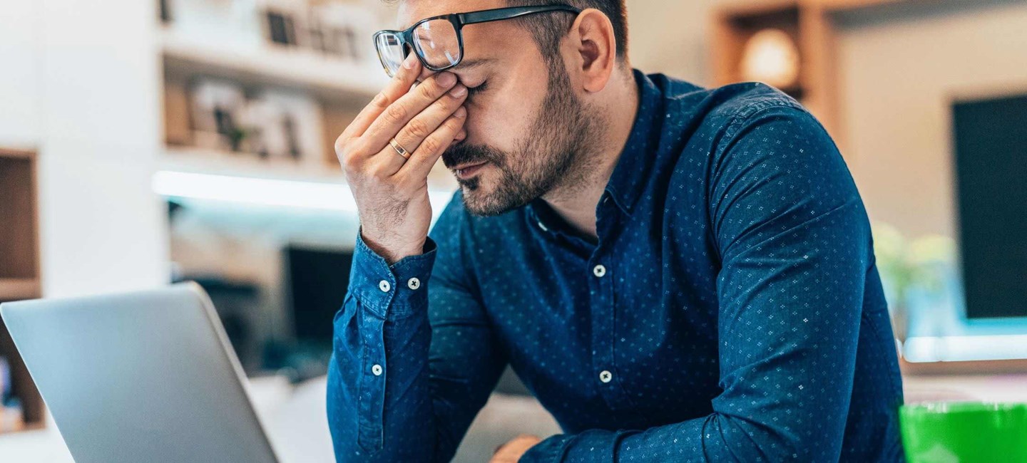 A man sat down in front of a laptop with his hand on his face, eyes closed