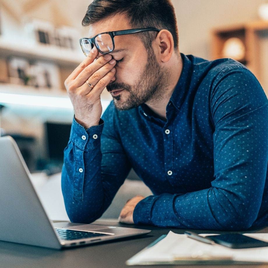 A man sat down in front of a laptop with his hand on his face, eyes closed