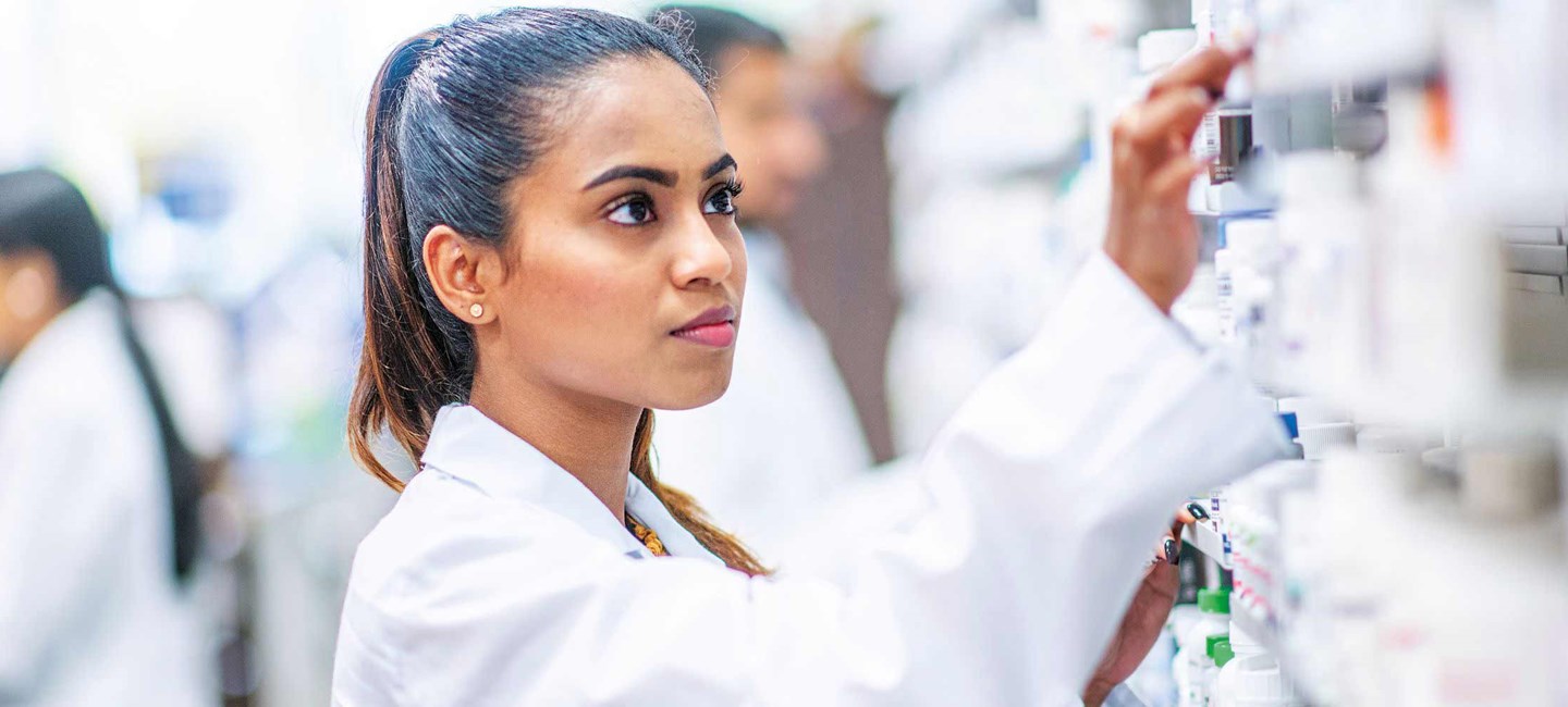 Woman in white lab coat reaching for a bottle of pills on a shelf