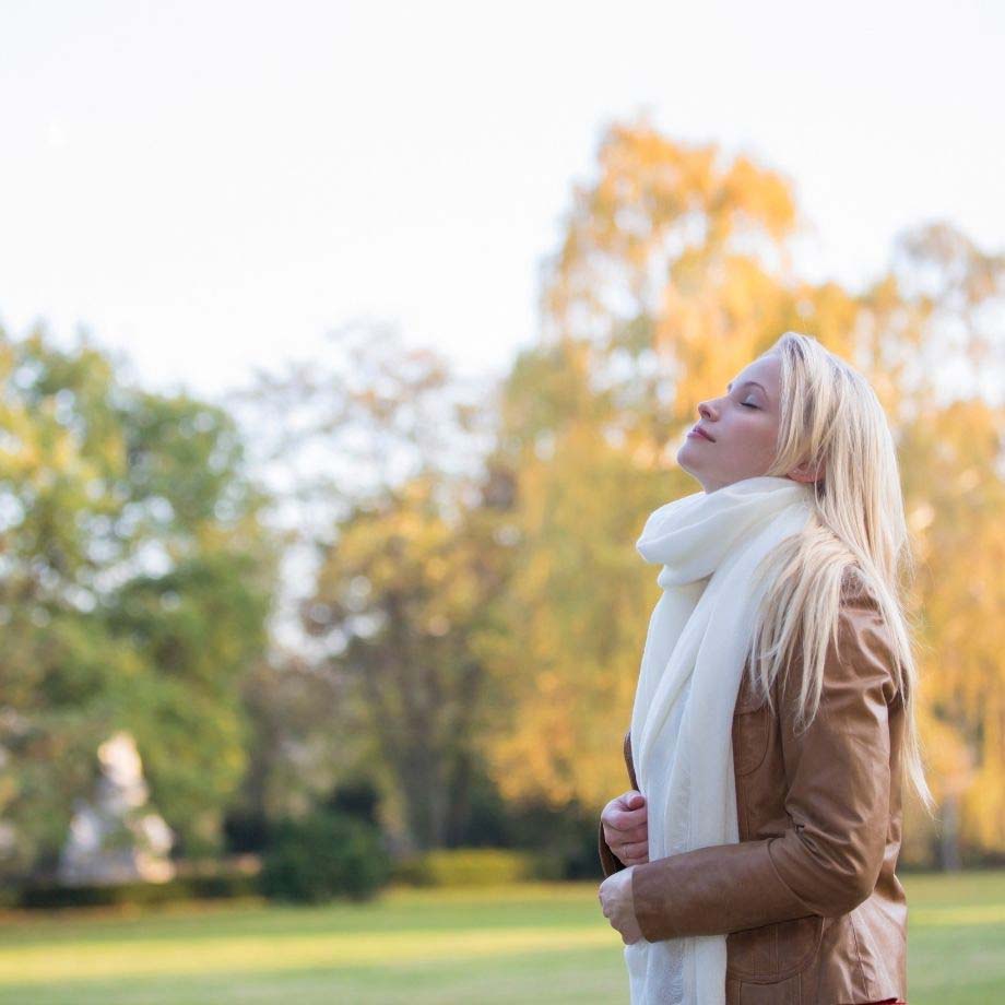 Woman in brown jacket, white scarf standing stationary with eyes closed and face slightly raised to sky